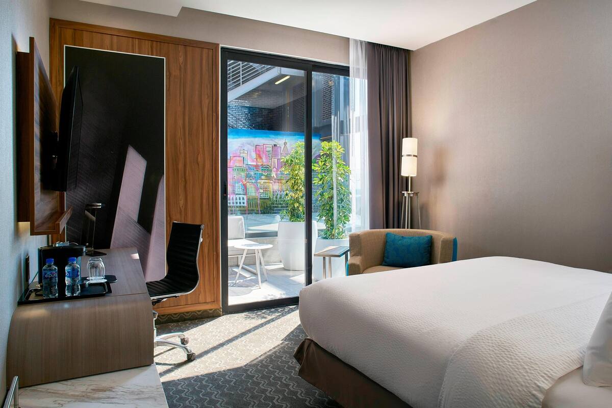 Une chambre d’hôtel avec un grand lit, un bureau avec un ordinateur, une chaise et une vue sur le paysage urbain, visible à travers une porte vitrée.