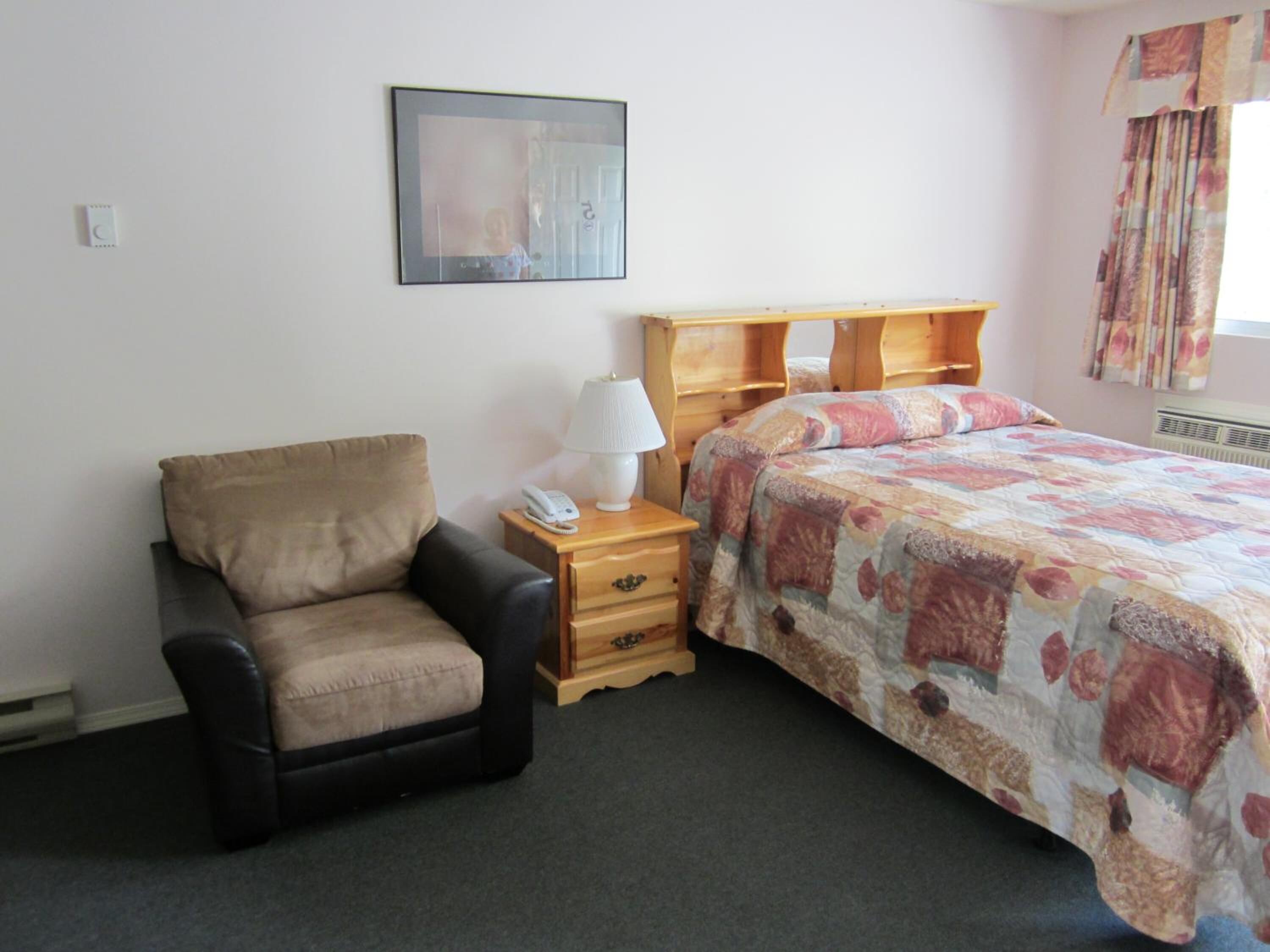 Une chambre à coucher avec un lit, une chaise, une table de chevet, une lampe et un tableau encadré au mur.