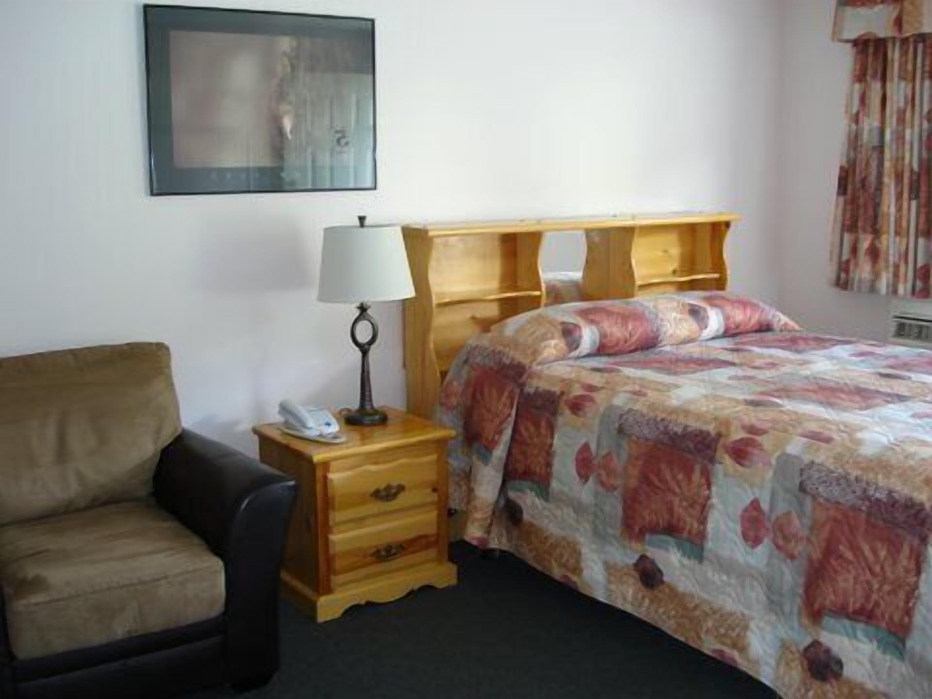 A bedroom with a bed, a chair, a nightstand with a lamp, and a painting on the wall.