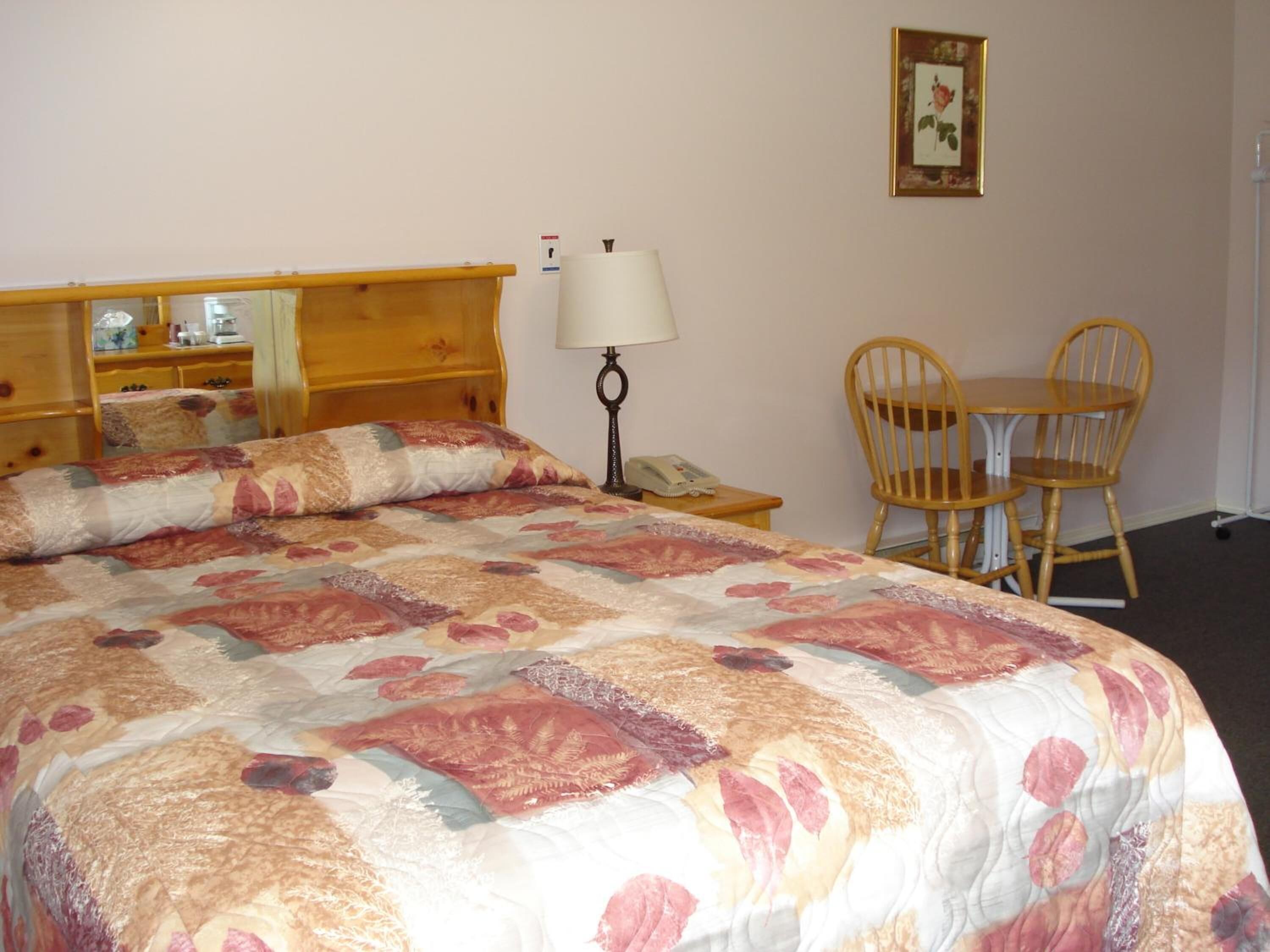 A hotel room with a large bed, a bedside table with a lamp, a chair, a small table, and a framed picture on the wall.