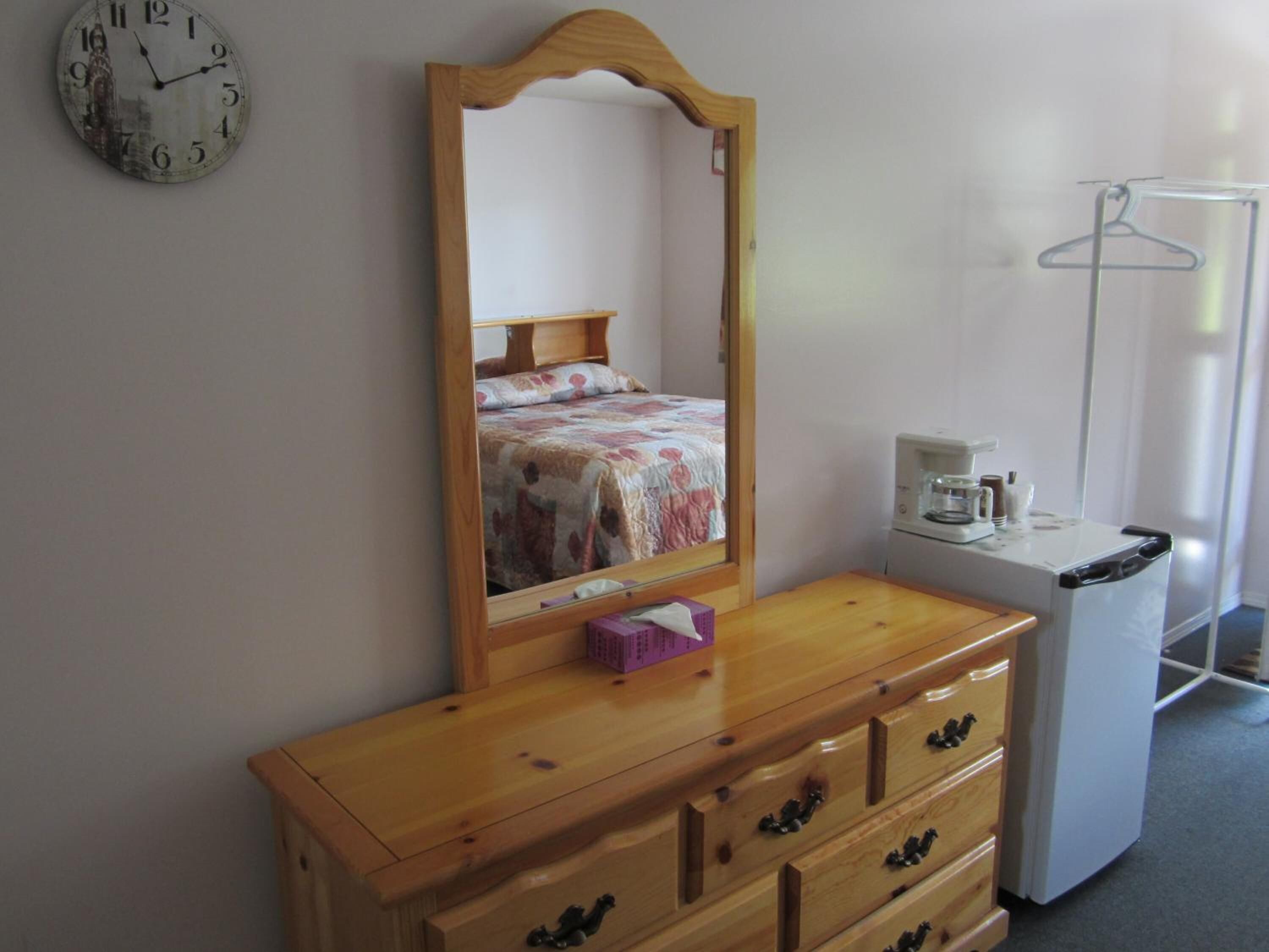 A bedroom with a wooden dresser, a mirror, a bed with a floral bedspread, a white refrigerator, and a wall clock.