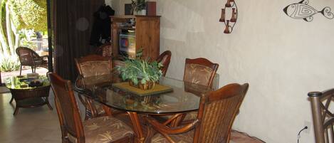 A dining area with a glass table surrounded by wicker chairs, a wooden cabinet with a plant, and a wall-mounted light fixture.