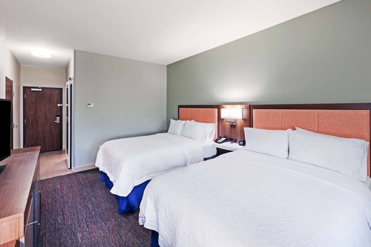 A hotel room with two beds, a desk, and a nightstand with a lamp.
