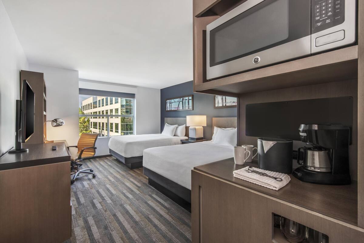 A hotel room with two beds, a desk, a microwave, a coffee maker, and a large window.