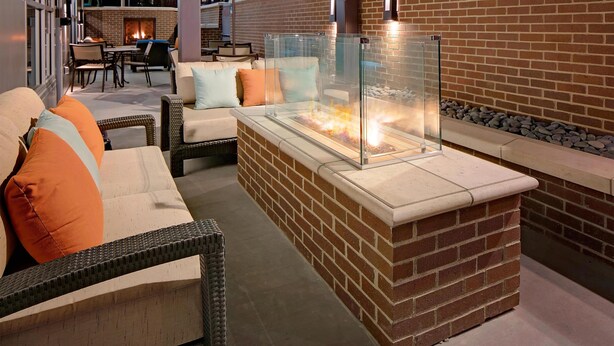 An outdoor seating area with wicker furniture, a glass-top fire feature, and a brick wall backdrop.