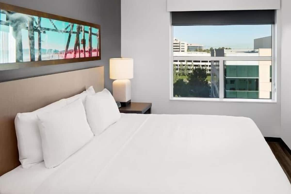 A hotel room with a bed, a nightstand, a lamp, a painting, a window, and a TV.