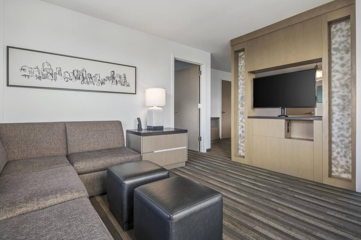 A hotel room with a sectional sofa, a flat-screen TV, a desk with a lamp, and a framed cityscape artwork.