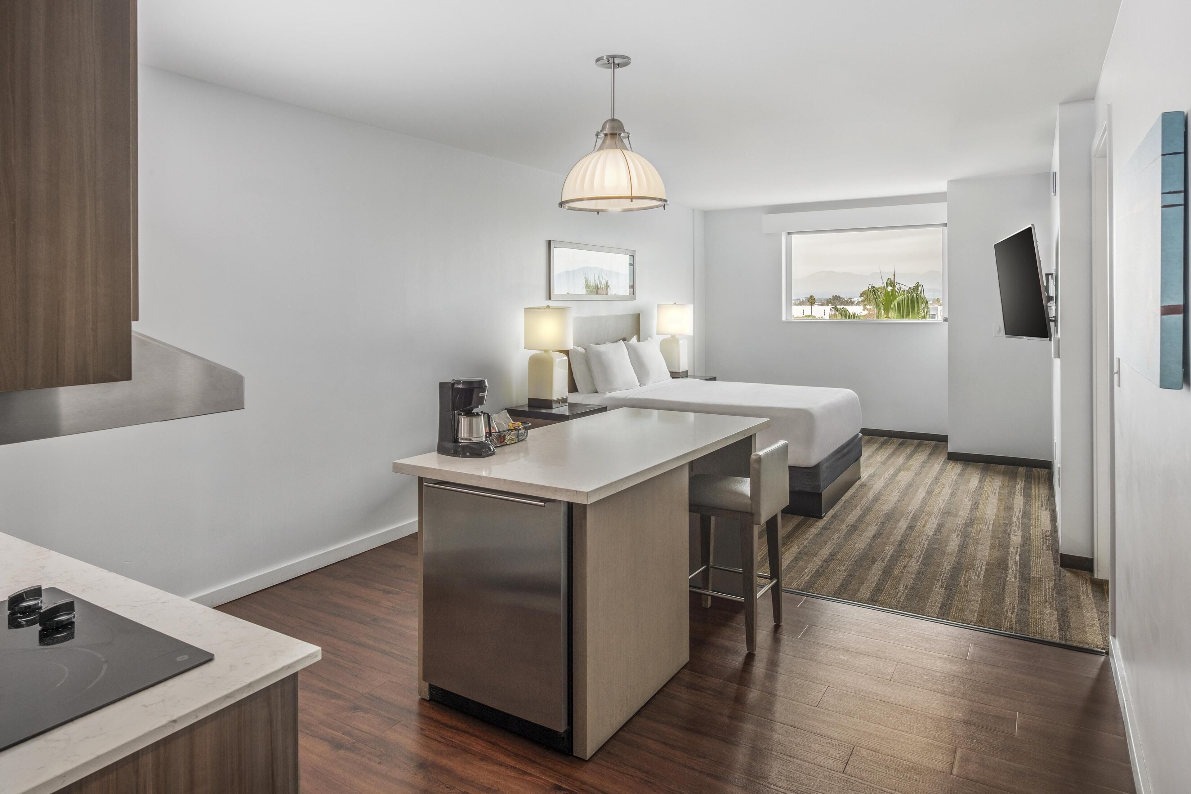 A modern hotel room with a kitchenette, a bed, a desk, and a large window offering a view.