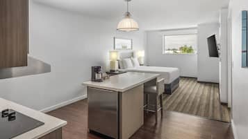 A modern hotel room with a kitchenette, a bed, a desk, and a large window offering a view.