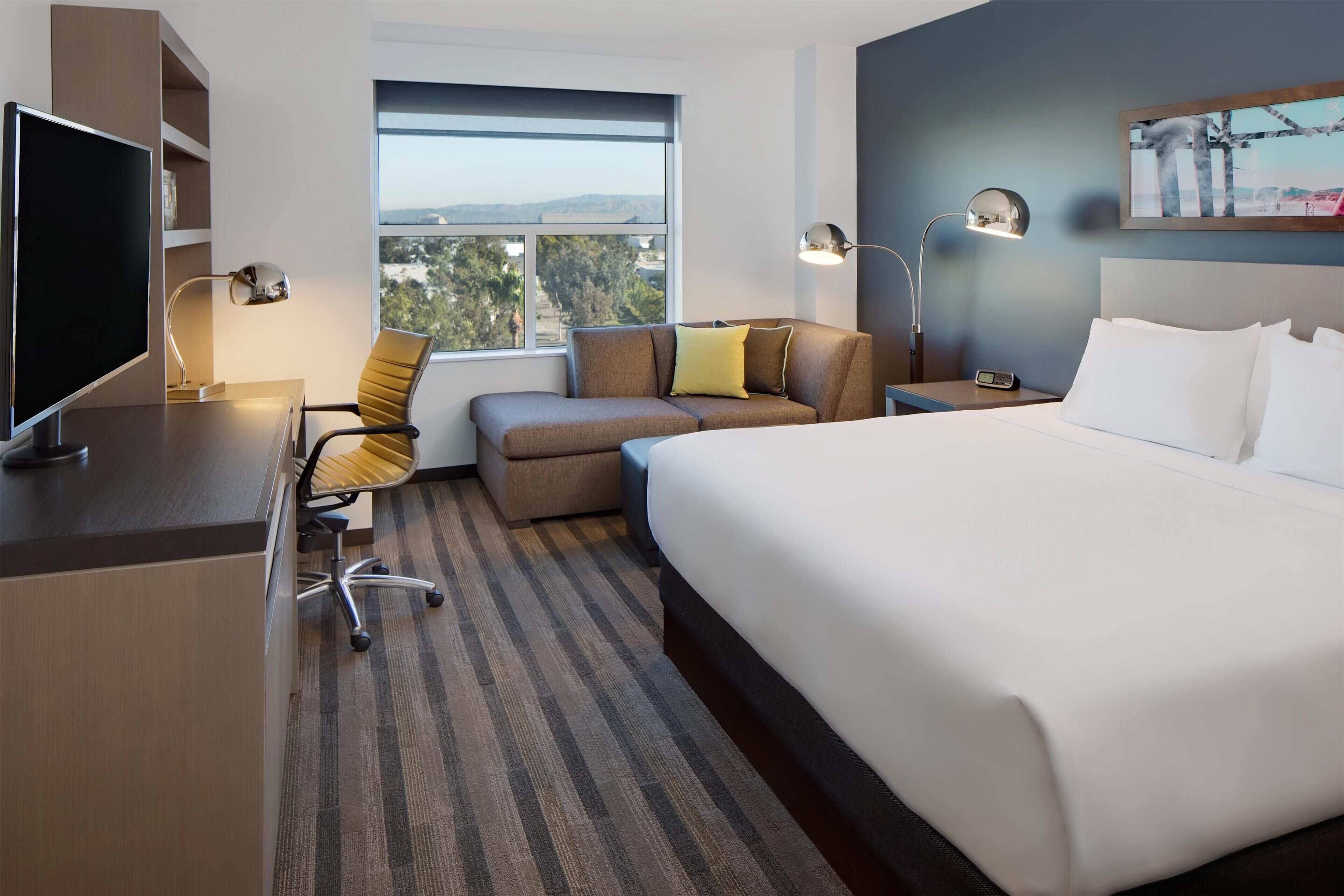 A hotel room with a bed, a desk with a computer, a chair, a sofa, and a window with a view.