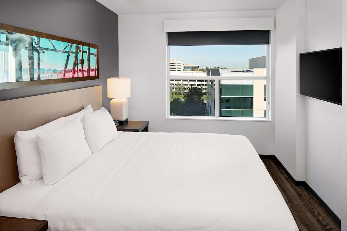 A hotel room with a large bed, a nightstand with a lamp, a TV, and a window with a city view.