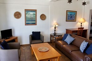 Un salón con un sofá de cuero marrón, una mesa de centro de madera, un sillón gris, un televisor sobre un mueble de madera y una mesa de comedor con sillas.