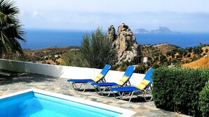 Una piscina con sillas azules, un solárium de piedra y vistas al mar y un paisaje rocoso.