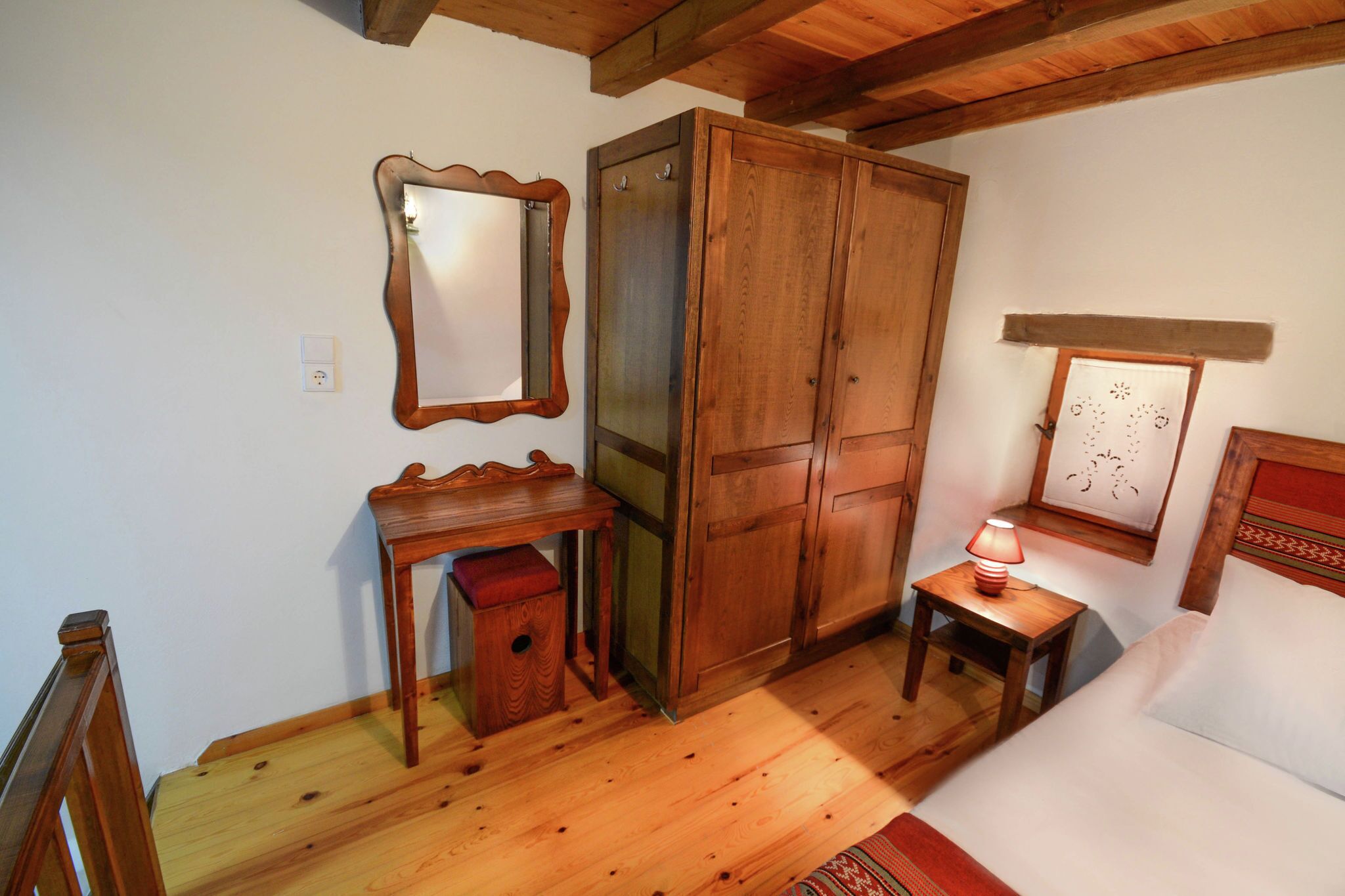 A room with a wooden wardrobe, a small table with a lamp, a mirror, and a bed.