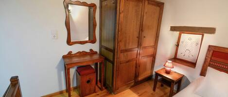A room with a wooden wardrobe, a small table with a lamp, a mirror, and a bed.