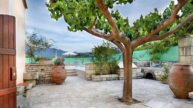 A stone-paved courtyard with a tree, large pots, and a pool area. - Villa in Crete with Vineyard Experience (Kalamitsi Alexandrou)