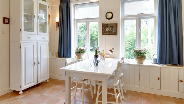 A dining area with a white table and chairs, a cabinet with glass doors, and large windows with blue curtains. - Holiday home in Krabbendam (Krabbendam)
