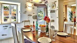 Un coin repas avec une table en bois dressée pour quatre personnes, des murs en pierre, un luminaire suspendu rouge et une vue par une fenêtre surmontée d’une arche en pierre.