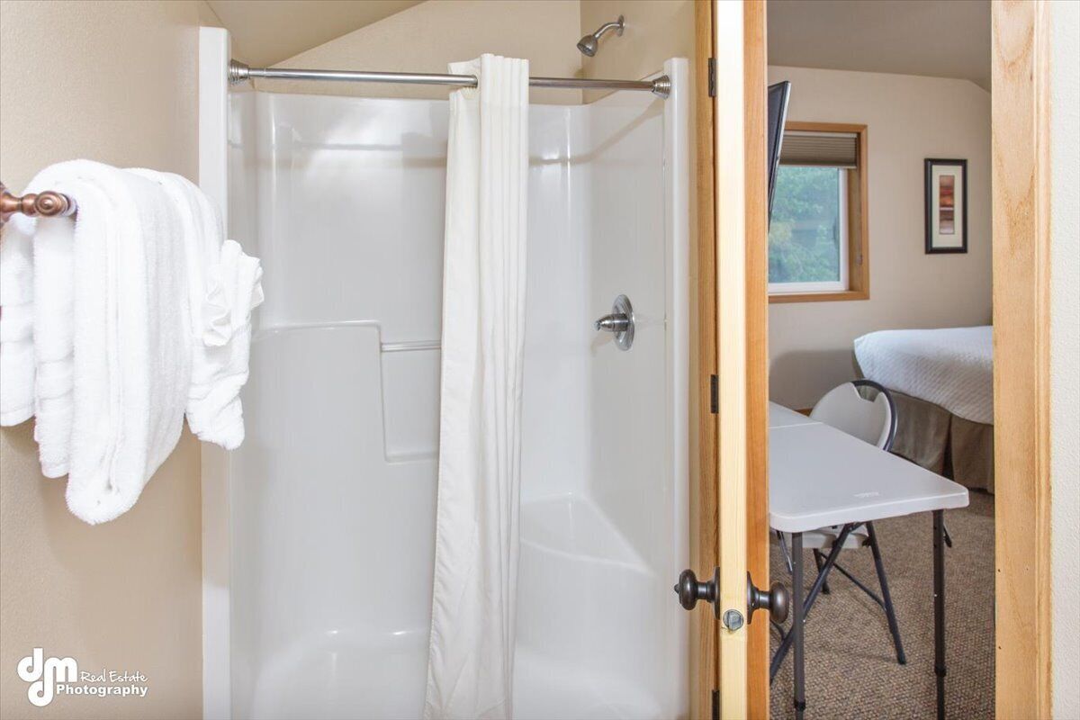 A shower with a glass door, a towel rack with white towels, and a small table with a chair in the adjacent room.