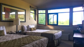 Habitación de hotel con dos camas, un escritorio, una silla, una lámpara y ventanas amplias con vistas a zonas verdes.