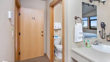 Ein Hotelbad mit einer Holztür, die die Nummer 14 zeigt, einer Toilette, einem Waschbecken mit Pflegeprodukten und einem Spiegel mit Handtuchhalter.