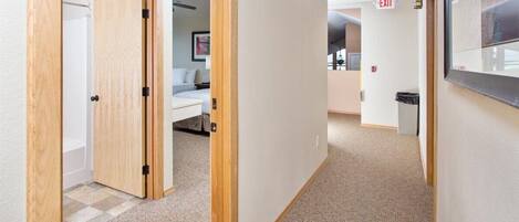 A hotel hallway with a wooden door, a bed in the background, and an exit sign.