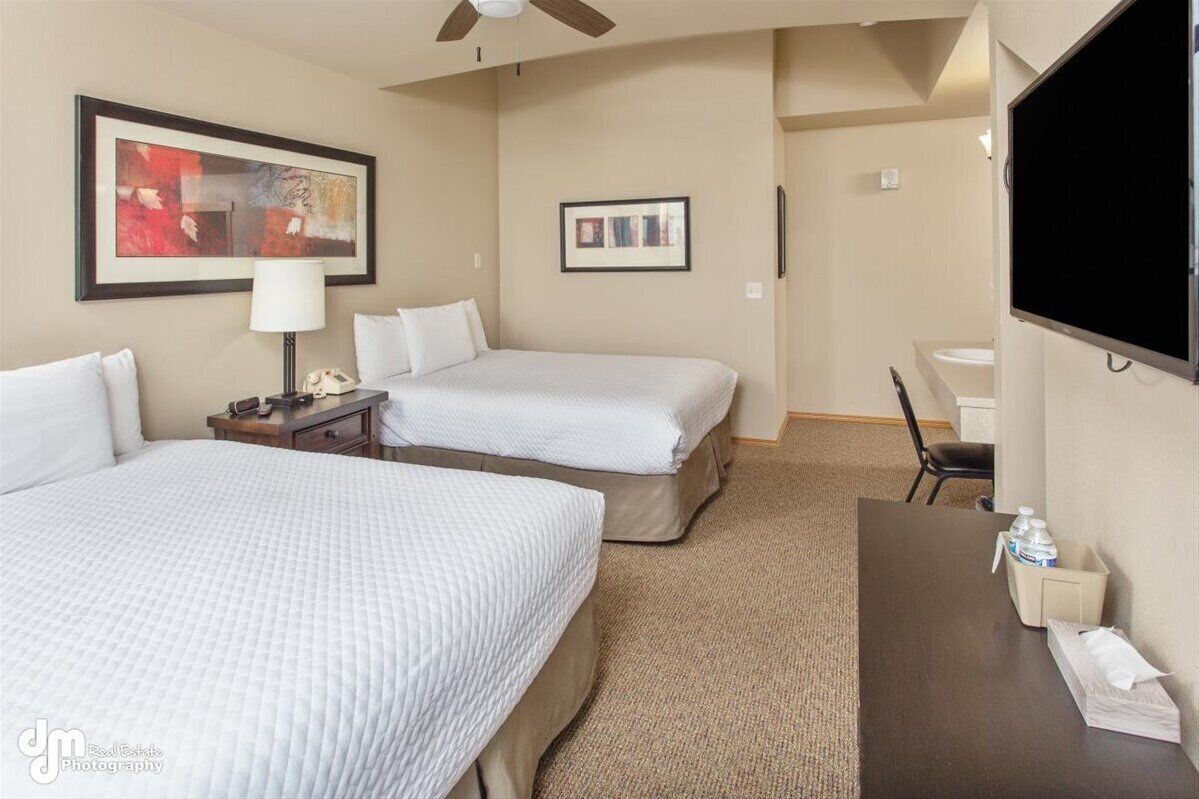 A hotel room with two beds, a desk, a chair, a TV, and a ceiling fan.