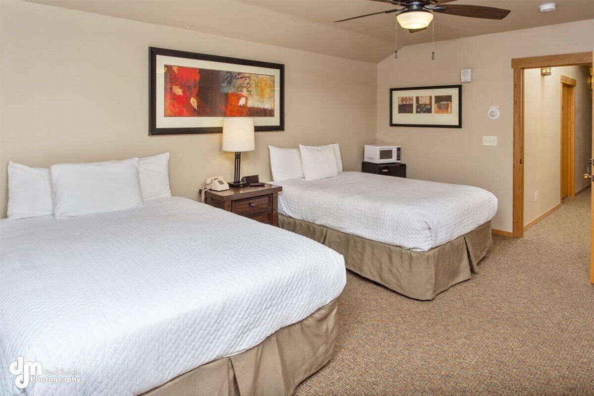 A hotel room with two beds, a ceiling fan, a nightstand with a lamp, and artwork on the walls.