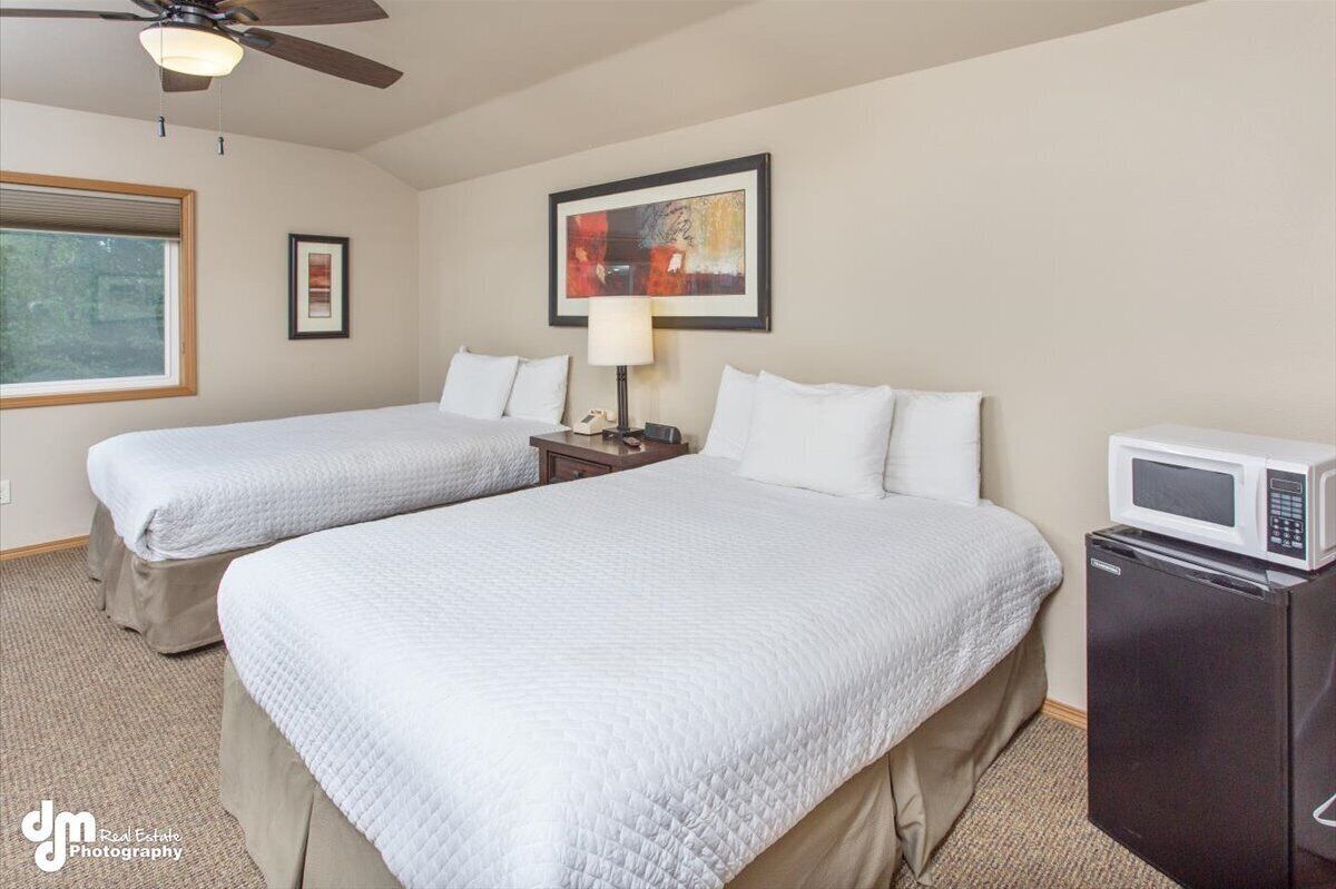 A hotel room with two beds, a ceiling fan, a painting, a lamp, and a microwave.