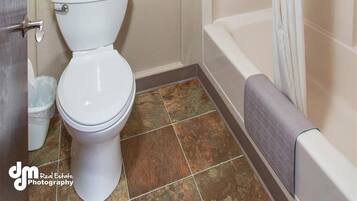 A bathroom with a toilet, a towel rack with a towel, a bathtub with a shower curtain, and a trash bin.