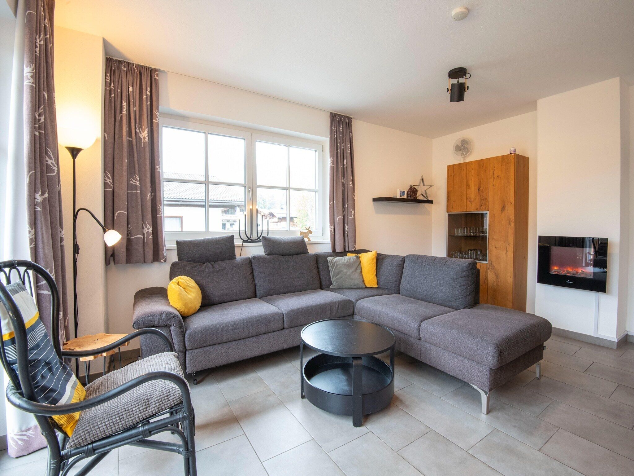 A modern living room with a grey sectional sofa, a round black coffee table, a black metal chair, and a wooden cabinet with shelves.