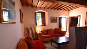 A cozy living room with a sofa, a coffee table, and a television. There are windows with curtains, a radiator, and framed pictures on the walls.