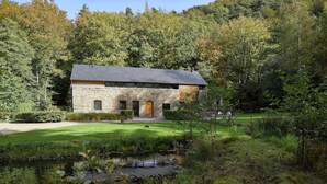 Une maison en pierre avec une porte et des fenêtres en bois, entourée d’un étang et de verdure.