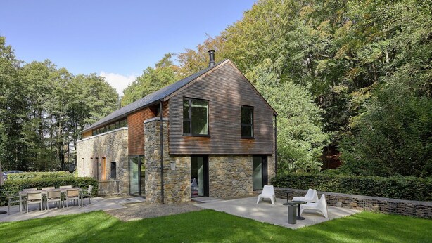 Une maison moderne avec une façade en pierre et en bois, un toit en pente et une pelouse bien entretenue agrémentée de mobilier d’extérieur.