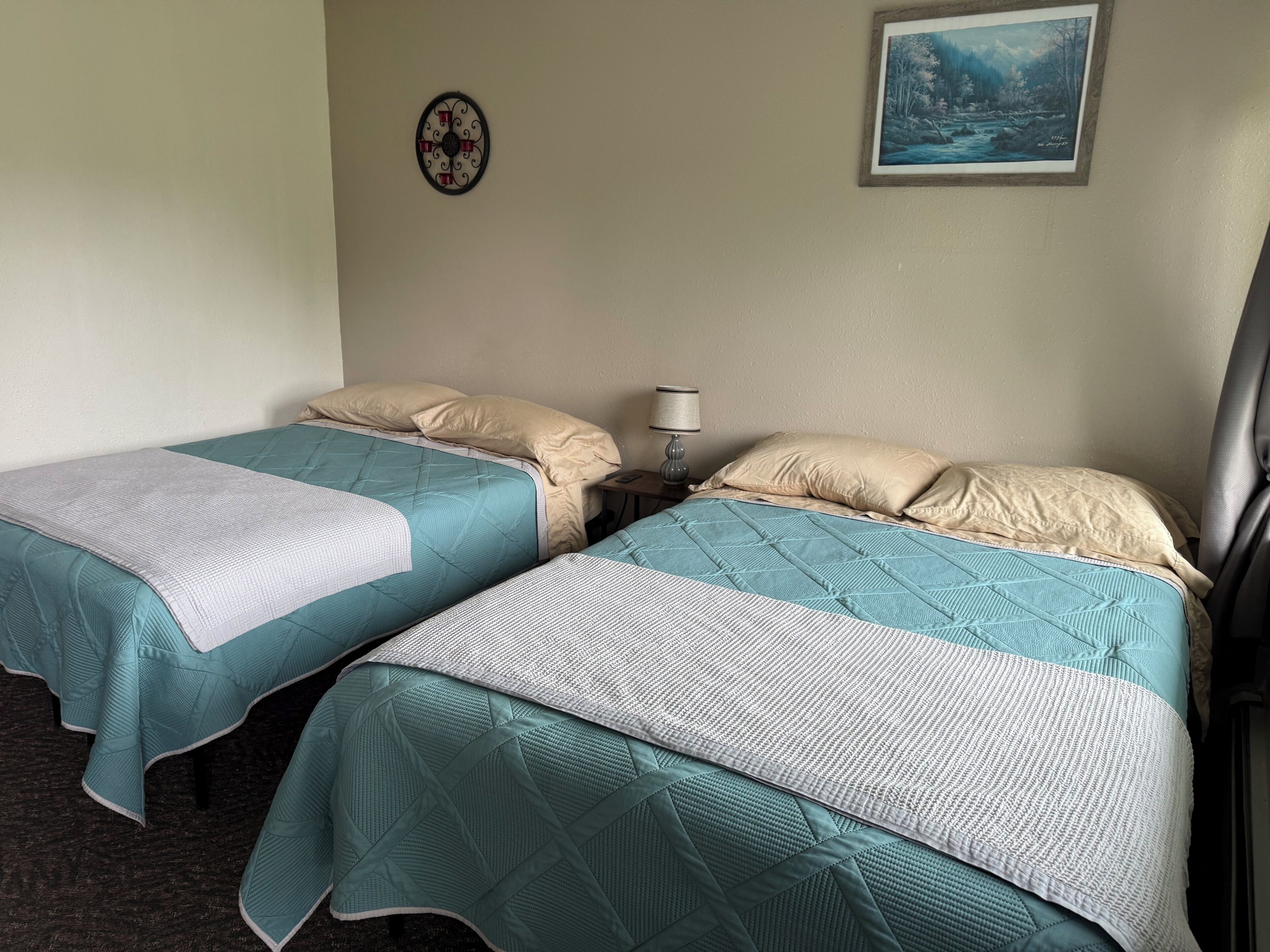 A hotel room with two beds, each with a teal bedspread and white pillows. There is a framed picture on the wall and a small table with a lamp.