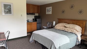 A compact hotel room with a bed, a small desk, a chair, a kitchenette, and a bathroom.