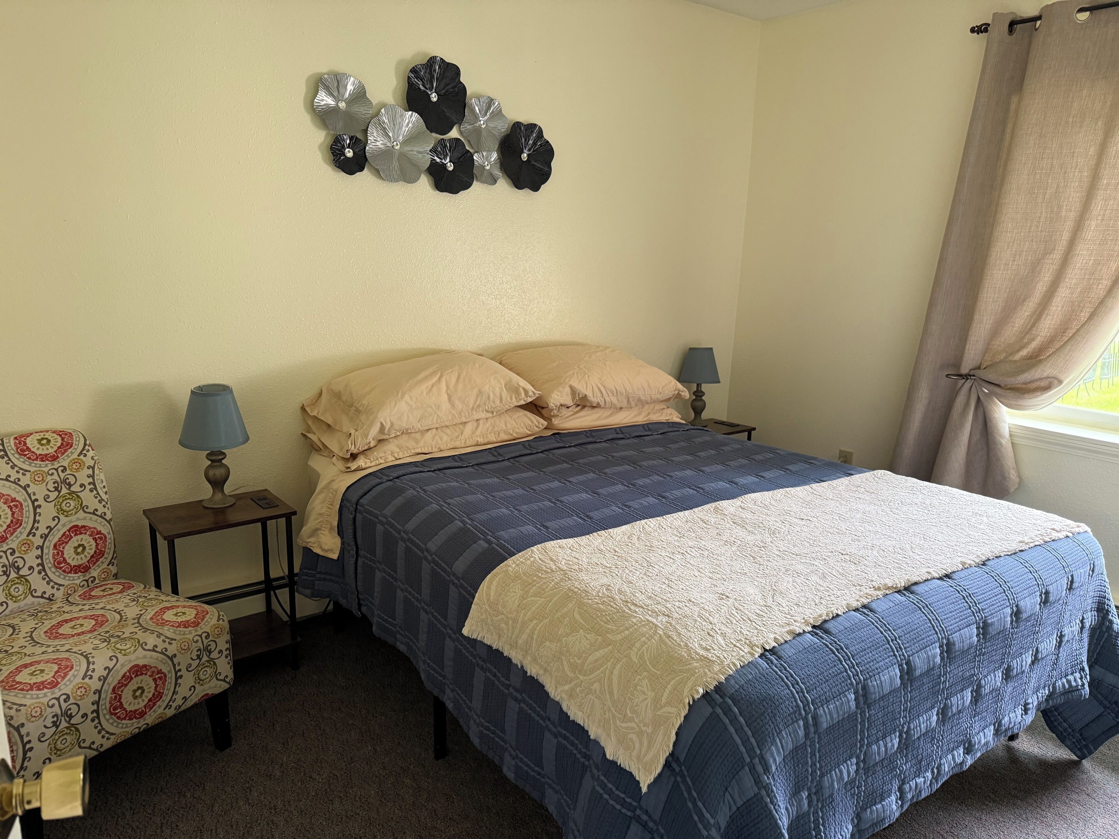 Un dormitorio con cama, dos mesitas de noche, lámpara, una silla y un adorno en la pared.