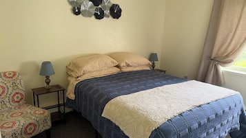 Un dormitorio con cama, dos mesitas de noche, lámpara, una silla y un adorno en la pared.