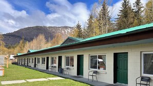 A row of single-story buildings with green doors and windows, a paved walkway, and a mountain backdrop. - Bird Creek Motel (Indian)