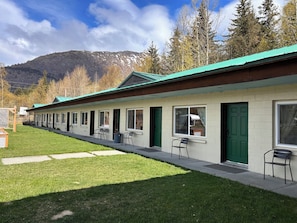 A row of single-story buildings with green doors and windows, a paved walkway, and a mountain backdrop. - Bird Creek Motel (Indian)
