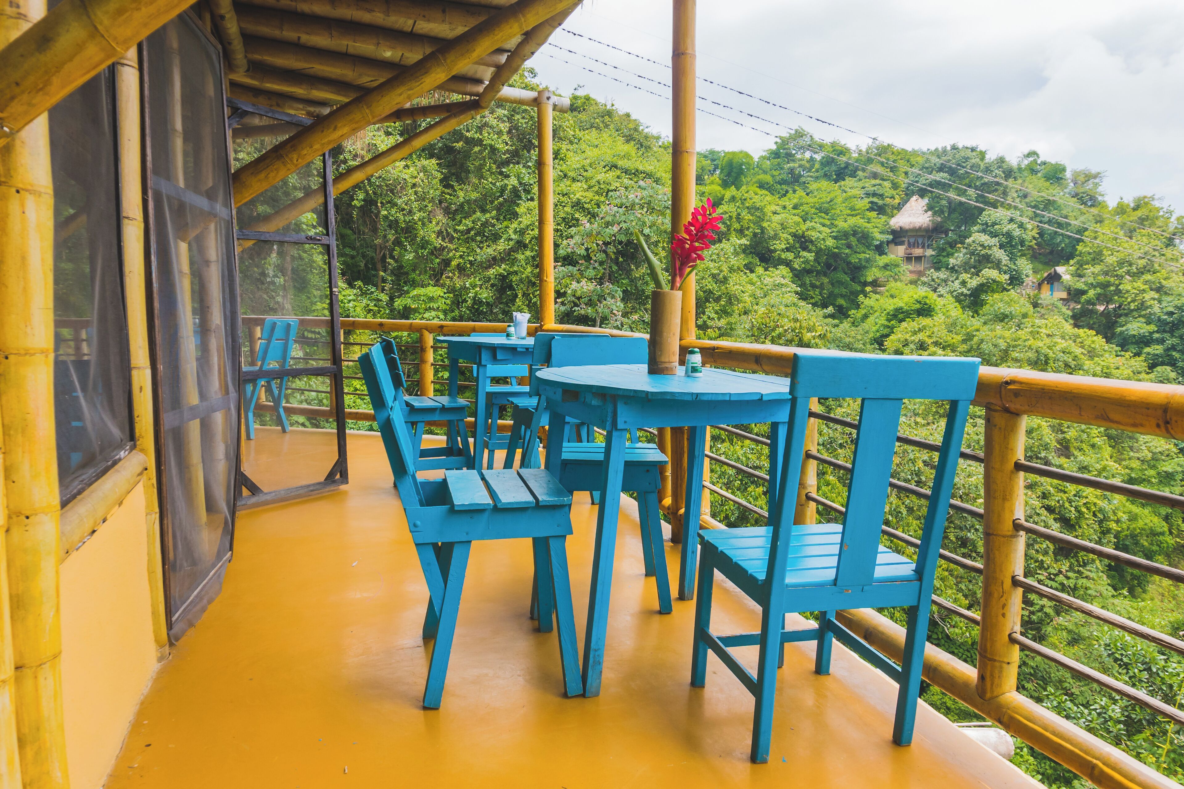 A covered outdoor seating area with blue chairs and tables, surrounded by a bamboo railing, overlooking a lush green landscape.