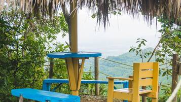 A rustic outdoor seating area with a wooden table and chair, surrounded by greenery and overlooking a mountainous landscape.