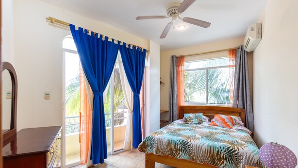 Un dormitorio con cama, ventilador de techo, ventana con cortinas, cómoda y mesita de noche con lámpara.