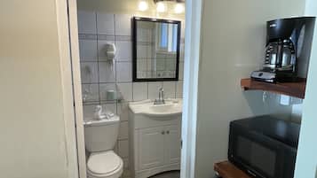 A compact bathroom with a toilet, sink, and a mirror above it. There is a hairdryer on the wall and a coffee maker on a shelf.