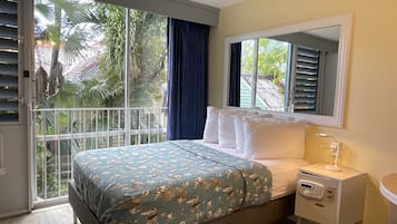 Een slaapkamer met een bed, een nachtkastje, een spiegel en uitzicht op palmbomen door een schuifglazen deur.
