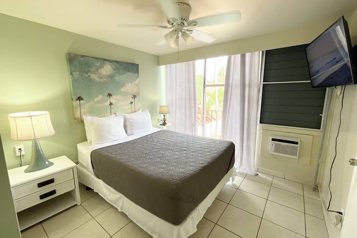 Un dormitorio con una cama, mesitas de noche, ventilador de techo, televisión y una ventana con cortinas.