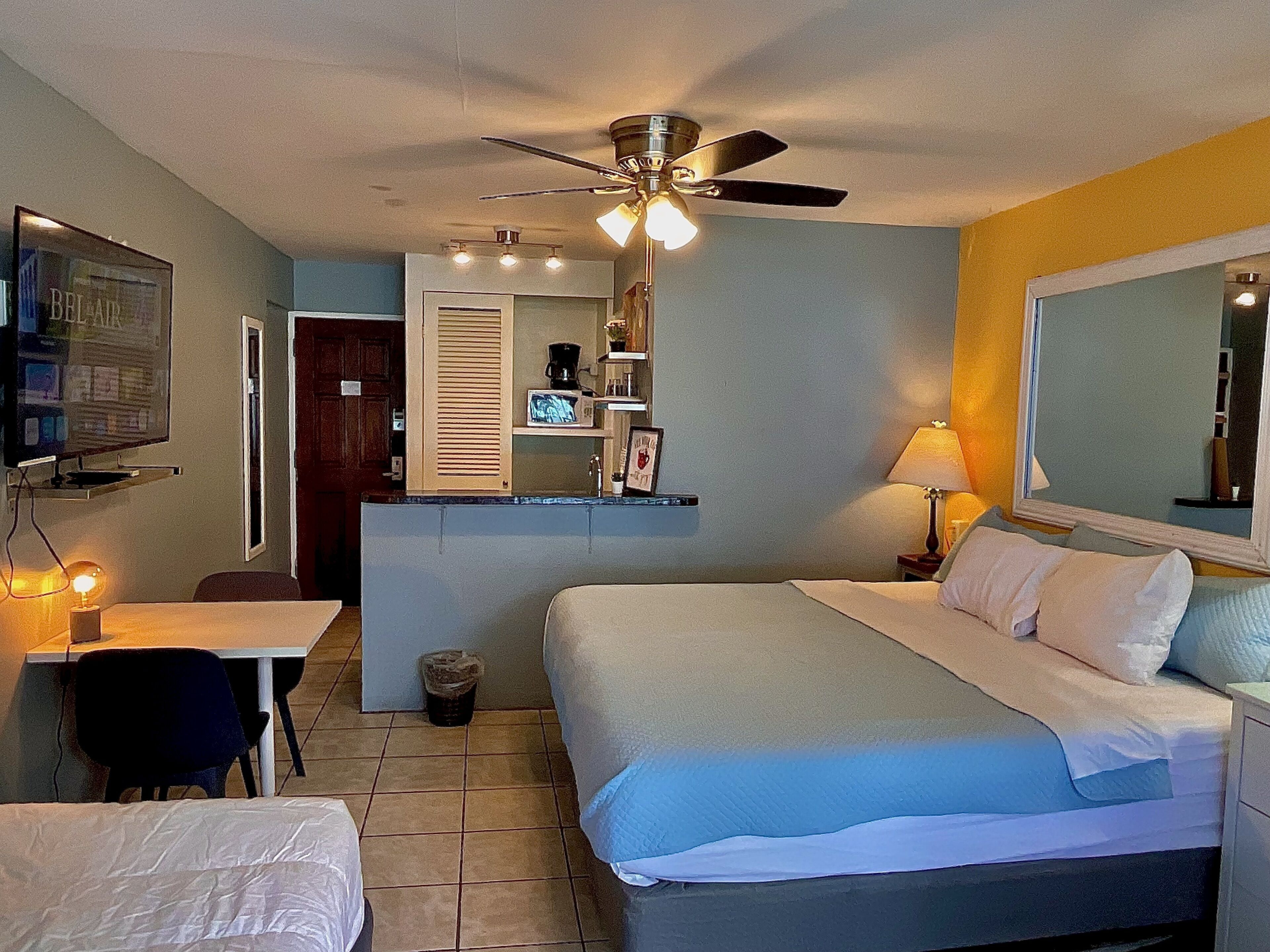 A hotel room with a bed, a desk, a chair, a ceiling fan, a mirror, and a small kitchenette.