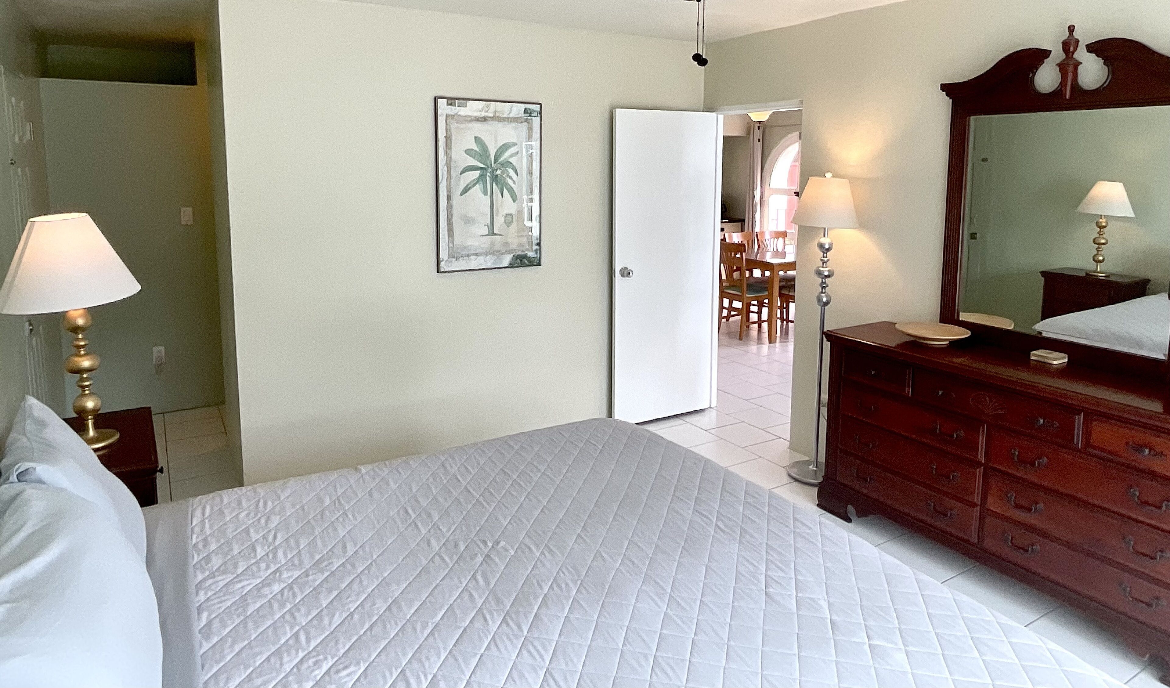 Habitación de hotel con cama, cómoda, espejo, lámpara y puerta que da a un comedor.