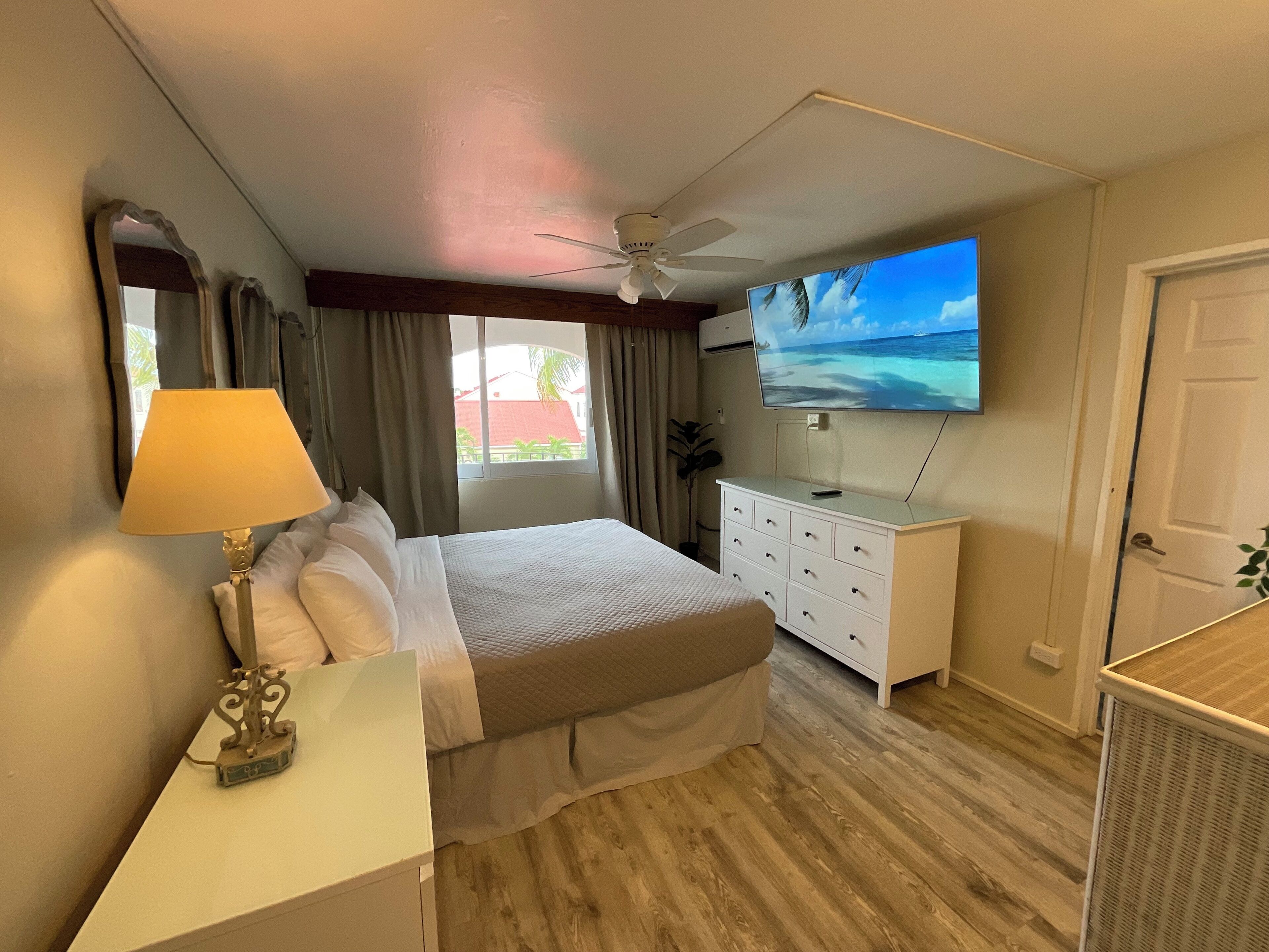 Une chambre d’hôtel avec un lit, un bureau doté d’une lampe, une commode, un téléviseur fixé au mur et un ventilateur de plafond.
