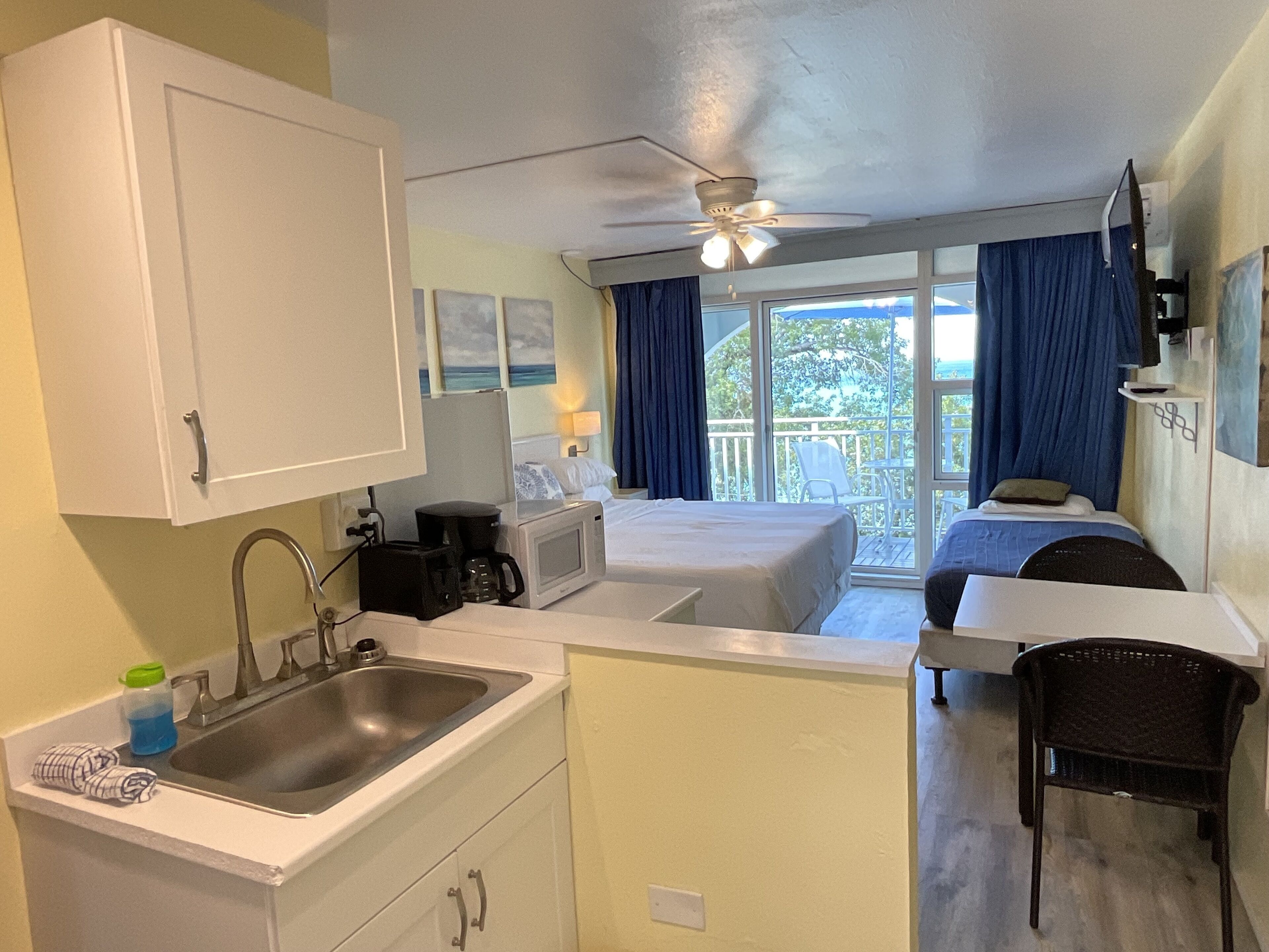 Une chambre d’hôtel compacte comprenant un coin cuisine, un lit, une petite table et une chaise. Elle dispose également d’un balcon avec vue sur l’extérieur.